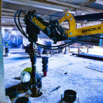 Worker operating a Brokk machine with Digga attachment for drilling in an industrial site.